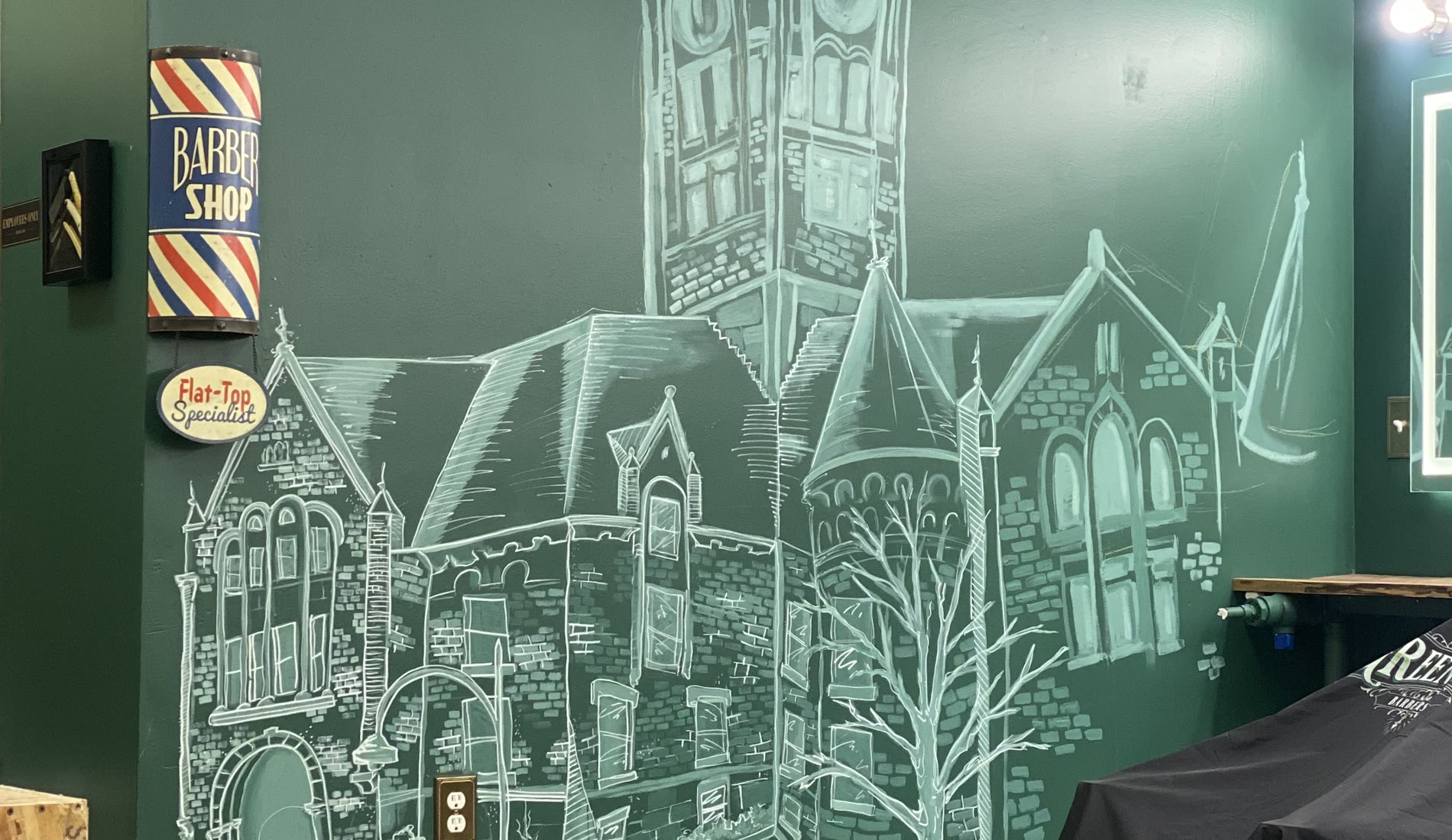 The hand-drawn chalk courthouse filling the shop wall, beside a vintage barber pole sign and a Flat-Top Specialist plaque
