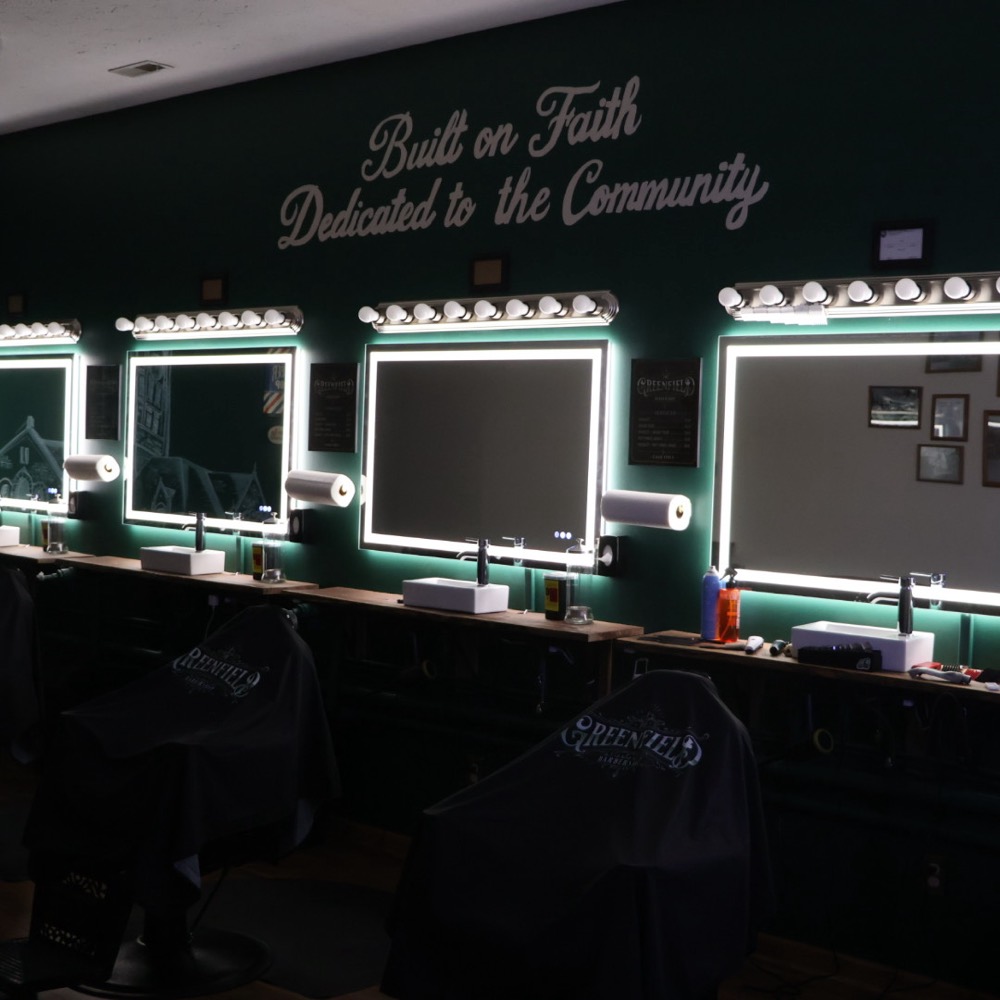The row of barber chairs at The Greenfield Barbershop lit by vanity bulbs against the deep green wall